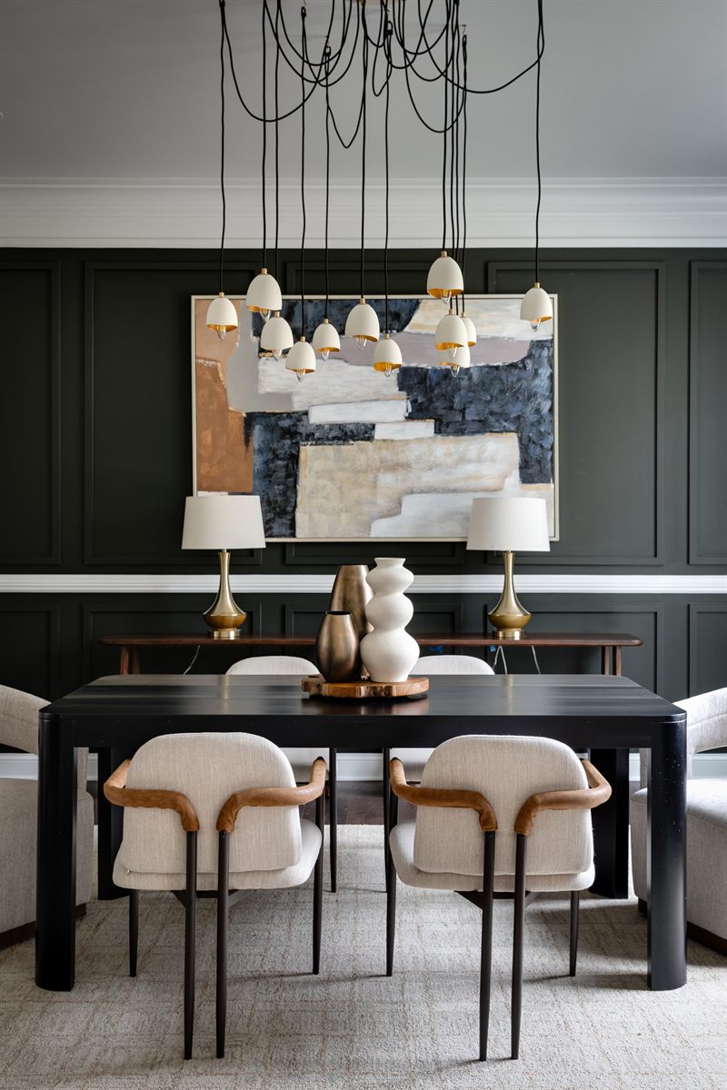 Staged dining room with a black table and cream armchairs against deep green panelled walls, under a cluster of pendant lights