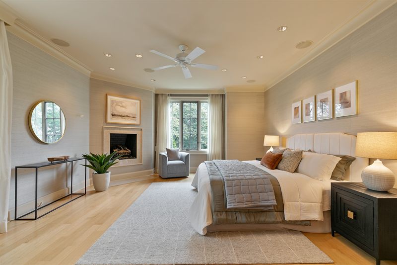 Primary bedroom with a fireplace, round gilt mirror, upholstered bed and an armchair by the window