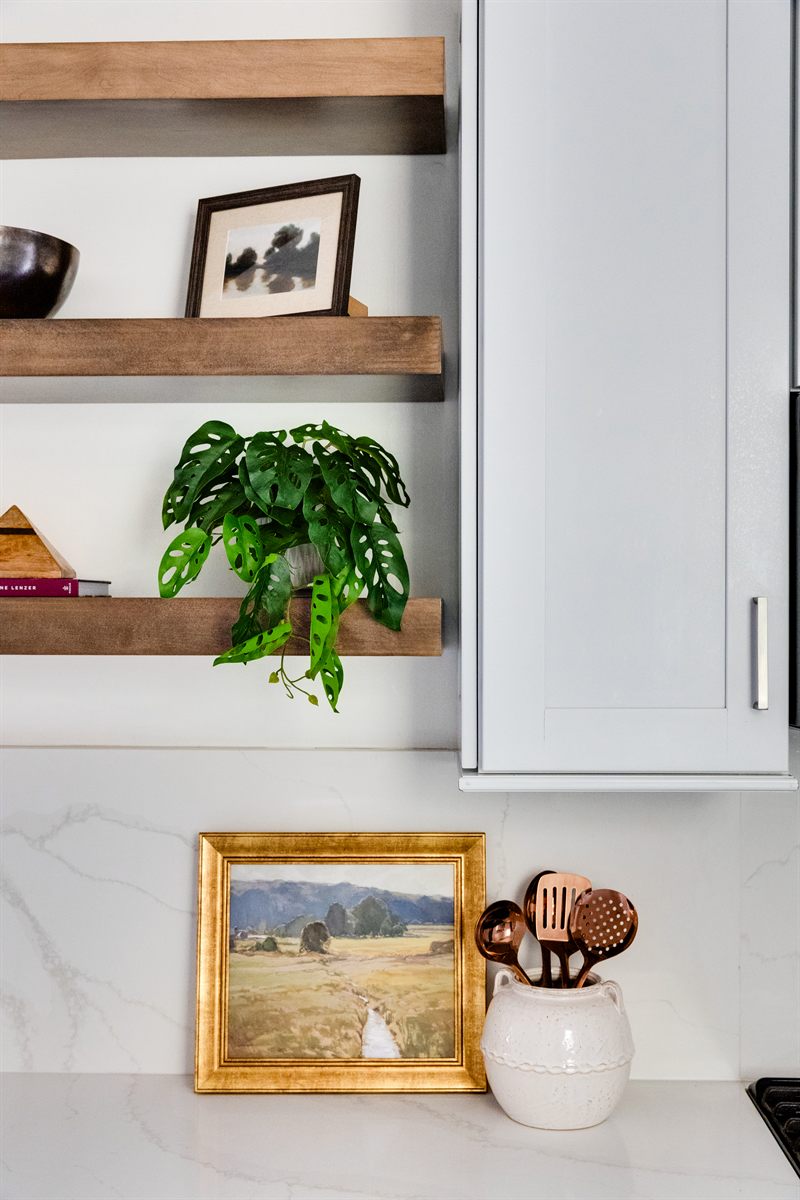 Wood floating shelves with a trailing plant above a marble counter, gilt landscape painting and utensil crock