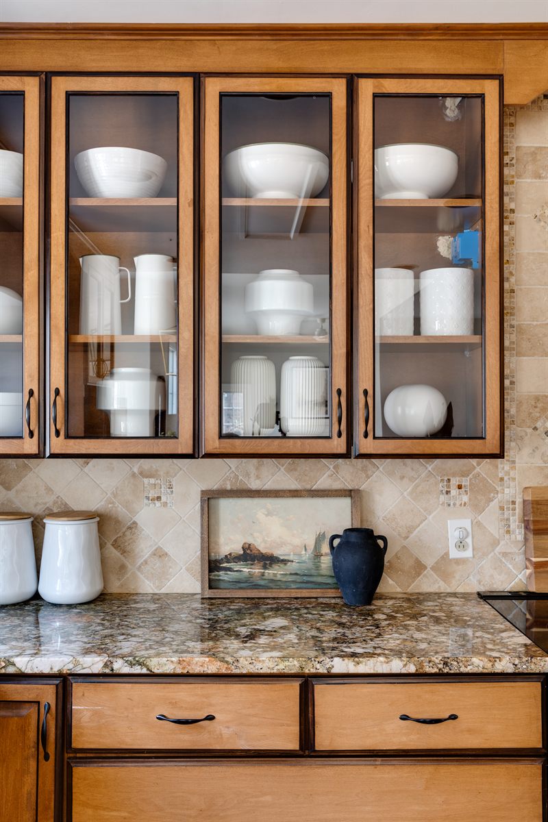 Glass-front cabinets filled with white serveware above a granite counter and a small seascape painting