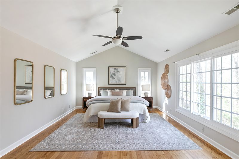 Primary bedroom with a vaulted ceiling, three gilt mirrors, upholstered bed and a bank of windows