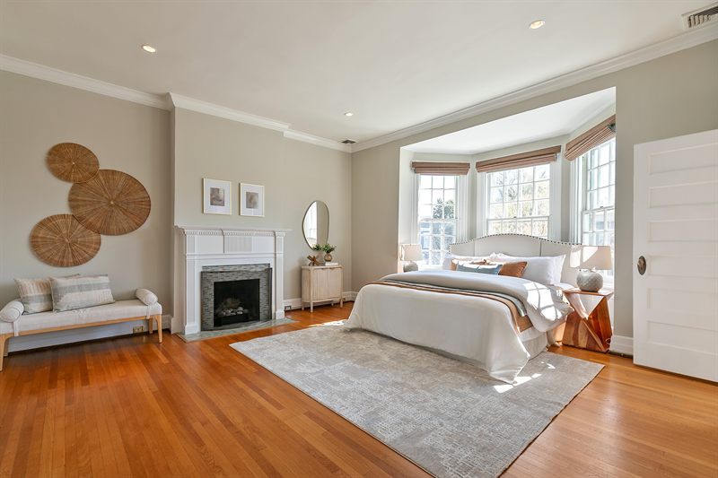 Primary bedroom with a marble fireplace, woven wall discs, a chaise and a bed in the bay window