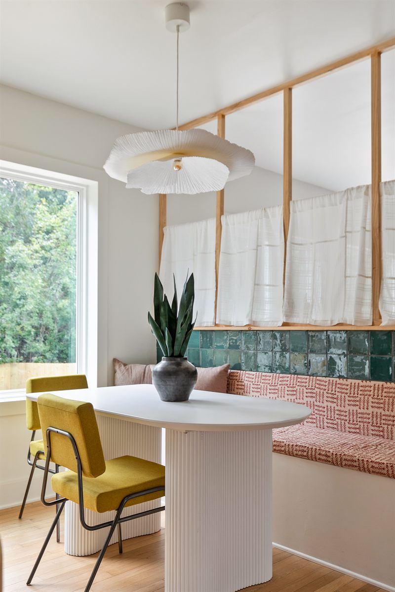 Fluted pedestal table with yellow chairs beside a green tiled banquette under a pleated pendant